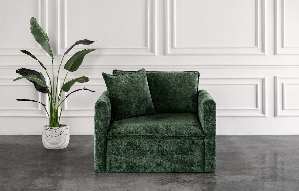 Green velvet armchair sofa bed with cushions in a room with white paneled walls and a plant.
