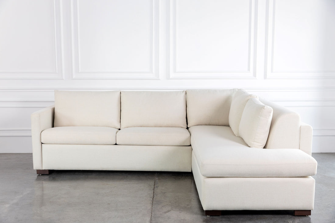 Beige sectional sofa bed in a minimalistic room with white walls and gray floor.